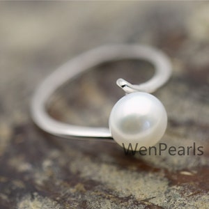 May include: A silver ring with a white pearl set on a curved band. The ring is on a brown wooden surface. The text "WenPearls" is visible in the image.