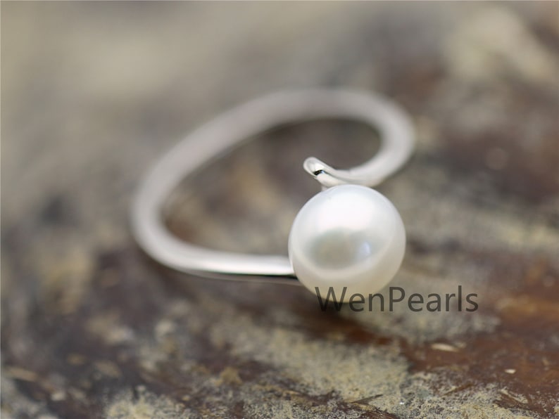May include: A silver ring with a white pearl set on a curved band. The ring is on a brown wooden surface. The text "WenPearls" is visible in the image.