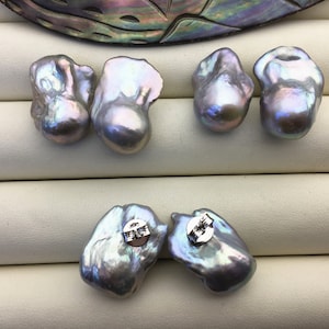 Puede incluir: Seis perlas grises iridiscentes de forma irregular. Dos están unidas a postes de aretes plateados. Las perlas tienen una superficie lisa y brillante con toques de azul y morado. Las perlas se exhiben sobre una superficie blanca.