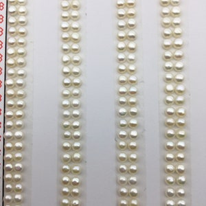 Puede incluir: Cuatro filas de cuentas de perlas blancas dispuestas verticalmente sobre un fondo blanco. Cada fila tiene aproximadamente 20 perlas.