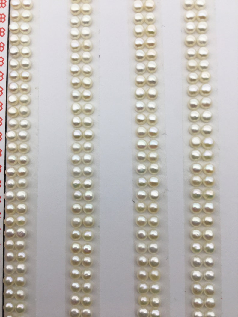 Puede incluir: Cuatro filas de cuentas de perlas blancas dispuestas verticalmente sobre un fondo blanco. Cada fila tiene aproximadamente 20 perlas.