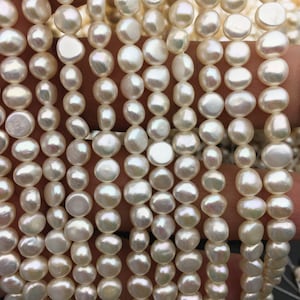 Puede incluir: Una hebra de perlas de agua dulce blancas, cada perla es redonda y lisa con un ligero brillo iridiscente. Las perlas están enhebradas en un hilo blanco y tienen un diámetro aproximado de 6-7 mm.