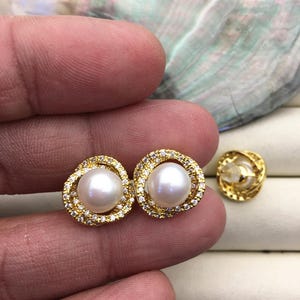 Puede incluir: Un par de pendientes de oro con una perla y pequeñas piedras transparentes engastadas en un diseño circular.