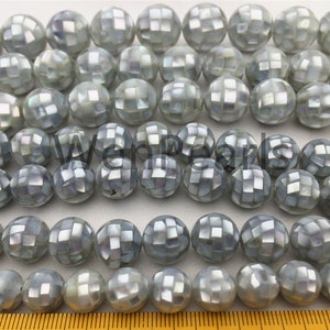 Könnte beinhalten: Eine Strähne runder, irisierender, grauer und weißer Muschelperlen mit einem facettierten, geometrischen Design. Die Perlen haben einen Durchmesser von etwa 10 mm.
