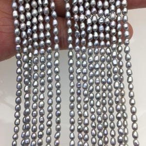 Könnte beinhalten: Stränge grauer Süßwasserperlen. Die oval geformten Perlen haben einen glänzenden Glanz. Die Perlen sind aneinandergereiht und erzeugen einen Kaskadeneffekt. Die Perlen sind neutral gefärbt und für verschiedene Schmuckdesigns geeignet.