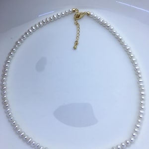 May include: A necklace composed of small, uniform white pearls strung together. The piece features a gold-toned metal lobster claw clasp and an adjustable chain extender with a small round bead at the end, completing the circular design.