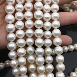 Könnte beinhalten: Eine Strähne weißer Süßwasserperlen, jede Perle ist rund und glatt. Die Perlen sind auf einem weißen Faden aufgereiht und in einer Reihe angeordnet. Die Perlen haben einen Durchmesser von etwa 8 mm.