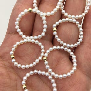 May include: Several delicate pearl rings with gold accents. Each ring is composed of small, round white pearls and a single gold bead. The rings are arranged in a cluster, showcasing their elegant design and potential for stacking or individual wear.
