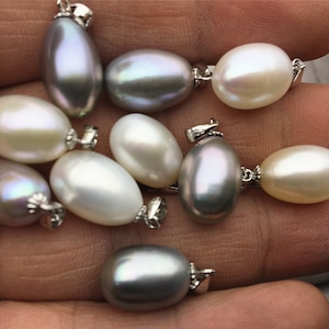 Puede incluir: Una colección de colgantes de perlas en varios tonos de blanco y gris. Los colgantes son de forma ovalada y tienen un acabado liso y brillante. Están enhebrados en cadenas de plata y están listos para ser usados como collares.