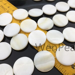 May include: A close-up of a collection of white round mother of pearl shell beads. The beads are arranged in a row on a yellow ruler with black markings. The ruler has the words "Made in China" printed on it.