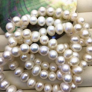 Può includere: Primo piano di più fili di perle bianche cremose e lucenti. Le perle sono di forma ovale e hanno una superficie liscia e riflettente. Lo sfondo presenta una conchiglia iridescente colorata, che aggiunge un tocco di bellezza naturale.