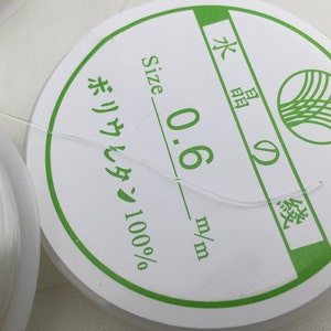 May include: A white spool of 0.6 mm nylon thread, labeled "Size 0.6 mm/m" and "ポリウレタン 100%" in Japanese. The spool is labeled with a green circle containing a stylized wave design.