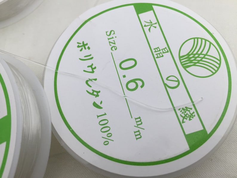 May include: A white spool of 0.6 mm nylon thread, labeled "Size 0.6 mm/m" and "ポリウレタン 100%" in Japanese. The spool is labeled with a green circle containing a stylized wave design.
