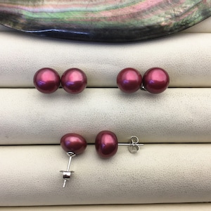 Puede incluir: Un par de pendientes con una perla burdeos en cada pendiente. Los pendientes están sobre una superficie blanca.