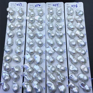 Puede incluir: Cuatro tarjetas de presentación blancas, cada una con diez pares de perlas blancas irregulares y brillantes. Las tarjetas tienen los números 053, 056, 055 y 056 escritos a mano en la parte superior, con filas numeradas del uno al diez.
