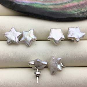 Può includere: Sei orecchini bianchi a forma di stella. Gli orecchini hanno una superficie strutturata e una lucentezza perlescente. Gli orecchini sono esposti su una superficie bianca, con una conchiglia scura e iridescente sullo sfondo. Gli orecchini sono a lobo.