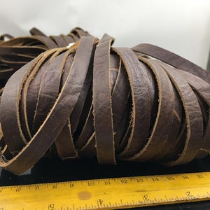 Peut inclure: Un lot de lanières de cuir brun, d'environ 30 cm de long, disposées en une bobine lâche. Le cuir est de couleur brun foncé et a une texture naturelle légèrement rugueuse.