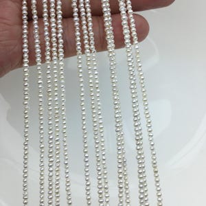 Puede incluir: Múltiples hebras de pequeñas perlas blancas. Las perlas son de tamaño uniforme y espaciadas, creando una apariencia refinada y elegante. Las hebras se muestran sobre un fondo claro, resaltando el brillo de las perlas.
