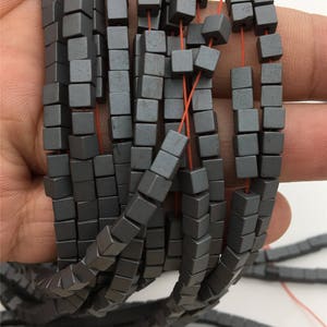 May include: A strand of gray hematite beads, each bead is a cube with slightly rounded edges. The beads are strung on a red thread.
