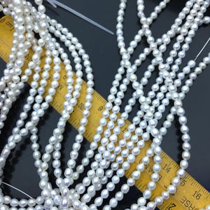 May include: Multiple strands of small, white, oval-shaped freshwater pearls are arranged over a yellow ruler. The ruler shows measurements in centimeters, with the pearls extending past the 16 cm mark on the scale.