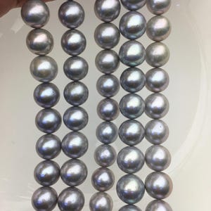 Puede incluir: Cuatro hilos verticales de perlas redondas, lustrosas y de color gris metálico. Cada hilo consta de múltiples perlas esféricas con una superficie lisa y reflectante, que muestran sutiles matices iridiscentes de azul y morado.