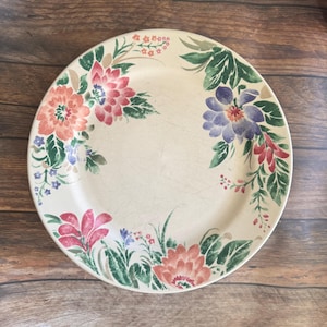 May include: Round, cream-colored plate with a floral design. The plate features a border of pink, red, purple, and orange flowers with green leaves. The plate is set against a wooden background.