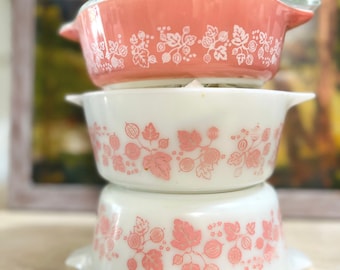 May include: A stack of three round glass bowls with handles. The top bowl is coral with a white gooseberry pattern. The middle and bottom bowls are white with a coral gooseberry pattern. Each bowl features a repeating design of leaves and berries.