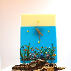 May include: A rectangular glass clock with a yellow top and blue lower section. The blue section features an underwater scene with colorful fish and plants. The clock hands are brown, and the clock is mounted on a piece of driftwood.