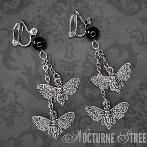 A pair of clip-on earrings lying on a grey damask background. Each earring has 2 Deaths Head Moth charms hanging by chains from a Black Onyx Bead.