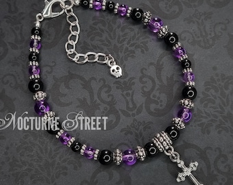 May include: A black and purple beaded bracelet with a silver skull charm and a silver cross pendant. The bracelet is on a dark patterned background.