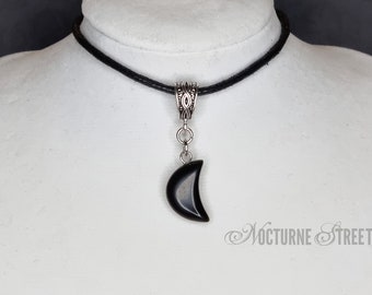 Puede incluir: Un collar gargantilla negro con un colgante de media luna plateado. El colgante está hecho de ónix negro y tiene una cadena plateada.