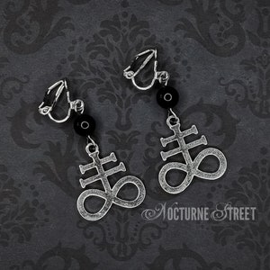 May include: A pair of silver clip-on earrings with black beads and a silver symbol of a cross with an infinity symbol. The earrings are on a black background with a floral pattern.