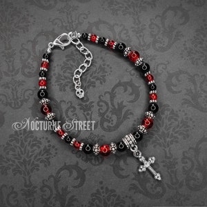 May include: Black and red beaded bracelet with a silver cross charm and a skull charm on the clasp. "Nocturne Street" is written on the background.