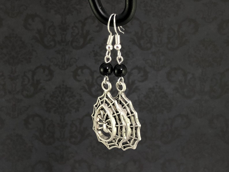 May include: Silver spider web earrings with black beads. The spider web design features a spider in the center.