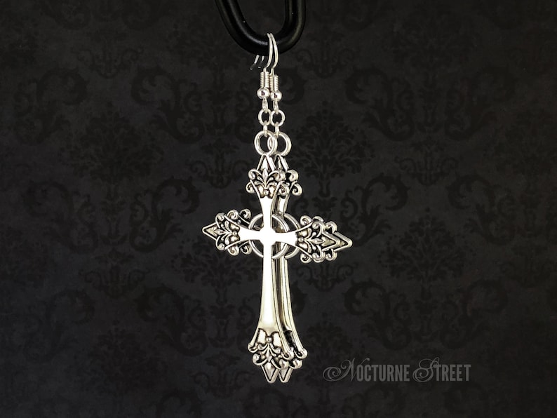 May include: Silver cross earrings with ornate details. The cross is double layered and has a delicate floral design.
