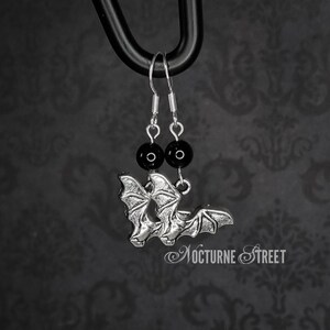 A pair of earrings hanging from a black hook in front of a grey damask background. The earrings each have a simple earring hook, a Black Onyx bead and a silver toned charm of a bat with it's wings spread.
