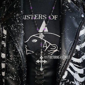 May include: A black leather jacket with white splatter paint and a black t-shirt with the text "Sisters of" and a graphic of a bear's head. A silver chain necklace with a cross pendant and purple beads hangs from the neck.
