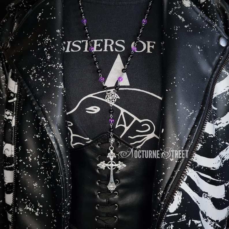 May include: A black leather jacket with white splatter paint and a black t-shirt with the text "Sisters of" and a graphic of a bear's head. A silver chain necklace with a cross pendant and purple beads hangs from the neck.