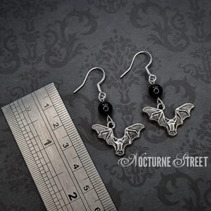 A pair of earrings laying on a grey damask background next to a ruler. The earrings each have a simple earring hook, a Black Onyx bead and a silver toned charm of a bat with it's wings spread. The ruler shows that the total length of the earrings is approximately 4.3cm