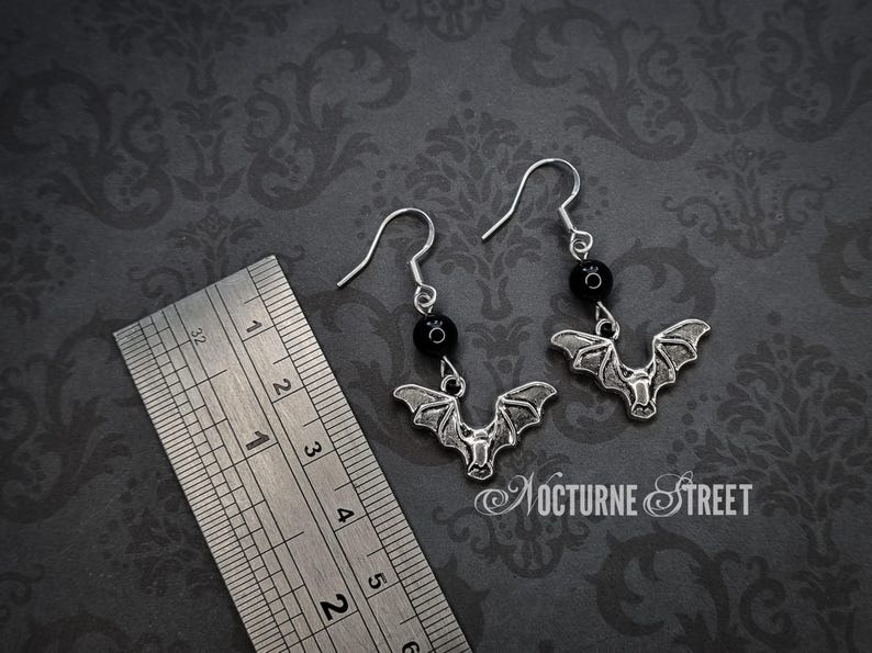 A pair of earrings laying on a grey damask background next to a ruler. The earrings each have a simple earring hook, a Black Onyx bead and a silver toned charm of a bat with it's wings spread. The ruler shows that the total length of the earrings is approximately 4.3cm