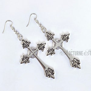 May include: A pair of silver cross earrings with intricate detailing. The crosses are ornate and have a pointed bottom.