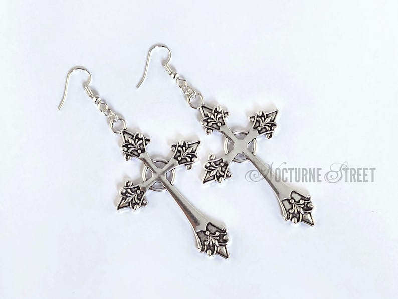 May include: A pair of silver cross earrings with intricate detailing. The crosses are ornate and have a pointed bottom.