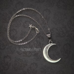 Silver crescent moon pendant, lying on a dark gothic damask background. The image shows the entire necklace with the chain, and the clasp has a decorative star.