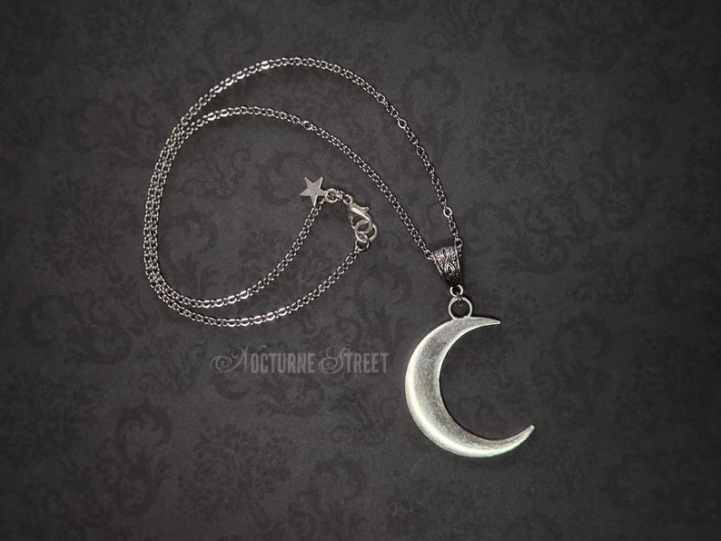 Silver crescent moon pendant, lying on a dark gothic damask background. The image shows the entire necklace with the chain, and the clasp has a decorative star.