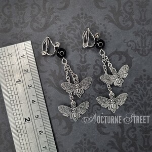 May include: A pair of silver clip-on earrings with a dangling chain and three silver moth charms. Each earring has a black bead at the top.