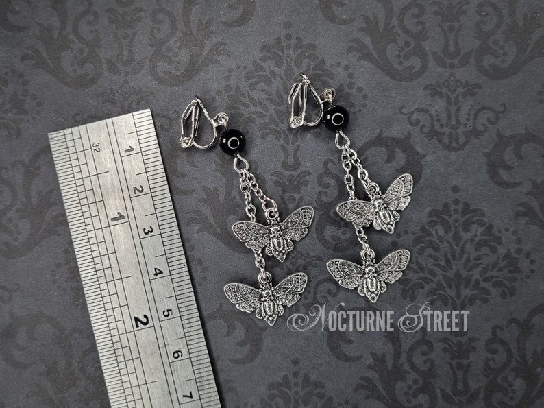 May include: A pair of silver clip-on earrings with a dangling chain and three silver moth charms. Each earring has a black bead at the top.