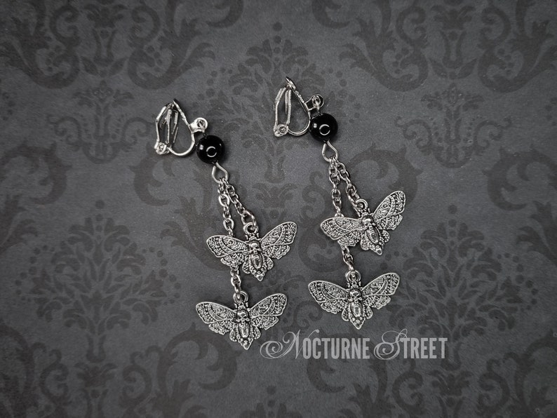 A pair of clip-on earrings lying on a grey damask background. Each earring has 2 Deaths Head Moth charms hanging by chains from a Black Onyx Bead.