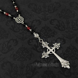 May include: A silver cross necklace with black and red beads. The cross has a detailed design and is attached to a chain with a circular pendant.