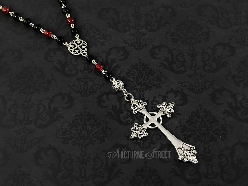 May include: A silver cross necklace with black and red beads. The cross has a detailed design and is attached to a chain with a circular pendant.