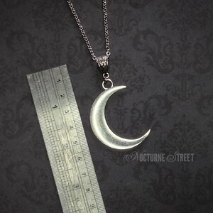 Silver crescent moon pendant lying on a dark gothic damask background next to a steel ruler. It shows the length of the moon and connector to measure approximately 6cm.